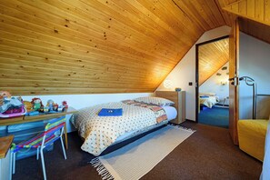 A bedroom with a sloped ceiling, a bed with patterned bedding, a desk with toys, a chair, and a door leading to another room.