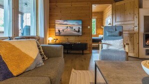 Een knus ingerichte kamer met een houten plafond, een grote flatscreen televisie aan de muur, een bank met een plaids en een fauteuil.