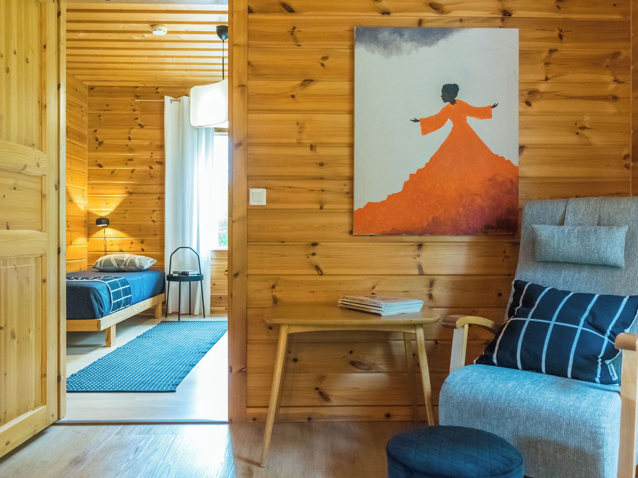 A wooden interior with a bed, a chair, a table, and a painting on the wall.