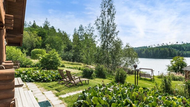 En træterrasse med udendørs møbler, en have med store, løvfyldte planter samt udsigt til sø og skov.