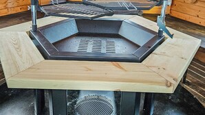 En sekskantet udendørs grill med metalrist, hvilende på metalstativer, placeret på en træplatform.