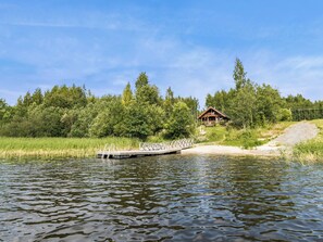 Puinen laituri, joka ulottuu järvelle, ja sen ympärillä on rehevää kasvillisuutta. Taustalla näkyy mökki.