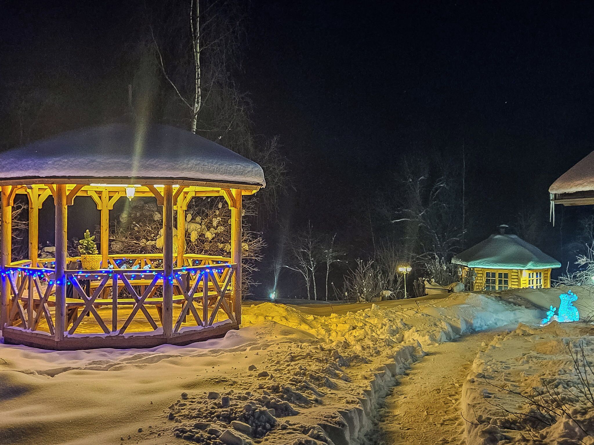 Zona nevada con un pabellón de madera iluminado por luces azules, un edificio pequeño al fondo y un sendero cubierto de nieve.
