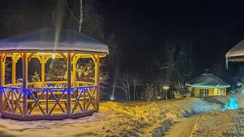 Zona nevada con un pabellón de madera iluminado por luces azules, un edificio pequeño al fondo y un sendero cubierto de nieve.