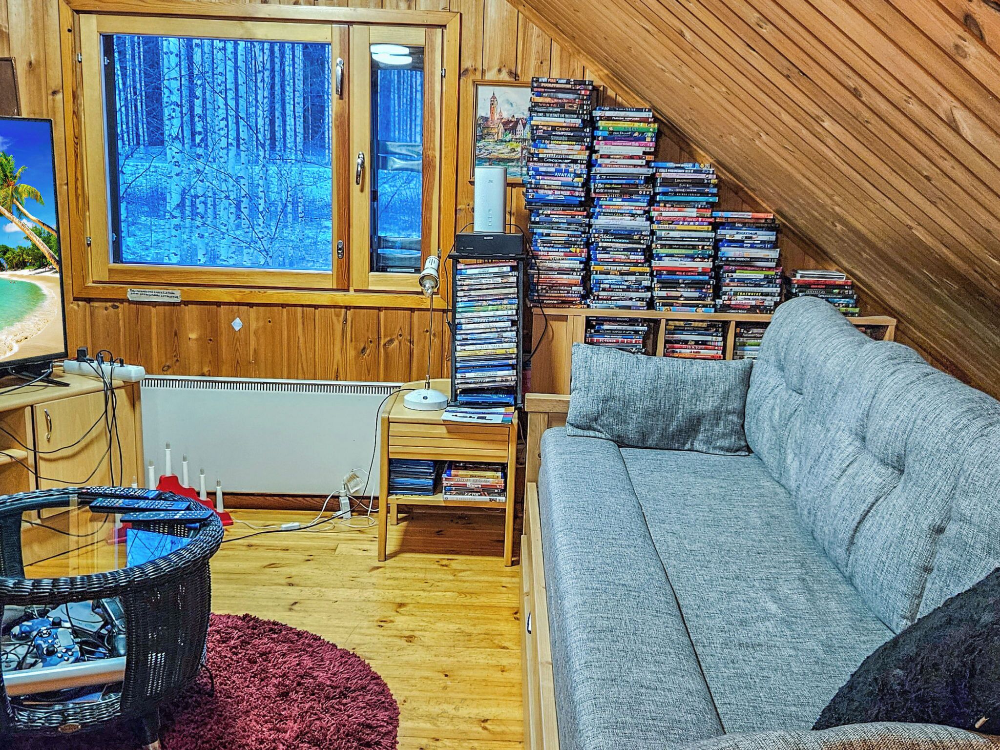 Tunnelmallinen huone, jossa on harmaa sohva, DVD-levyillä täytetty kirjahylly, ikkuna, josta avautuu näkymä metsään, sekä pieni pöytä, jossa on lamppu.