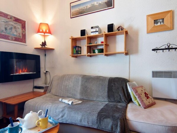 A cozy living room with a fireplace, a TV, a sofa, a coffee table with a teapot and cups, and shelves with books and decorative items.