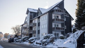 A multi-story residential building with balconies, a snow-covered landscape, and a clear sky. - Apartment - Im Hohlen Seifen 10-H | Winterberg (Winterberg)