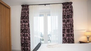 A hotel room with a bed, curtains, a nightstand, and a view of buildings through the window. - Apartment - Im Hohlen Seifen 10-H | Winterberg (Winterberg)
