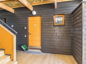 Een houten chalet met een deur waarop '18' staat, een klein raam en een houten trap naar een hoger gelegen verdieping.