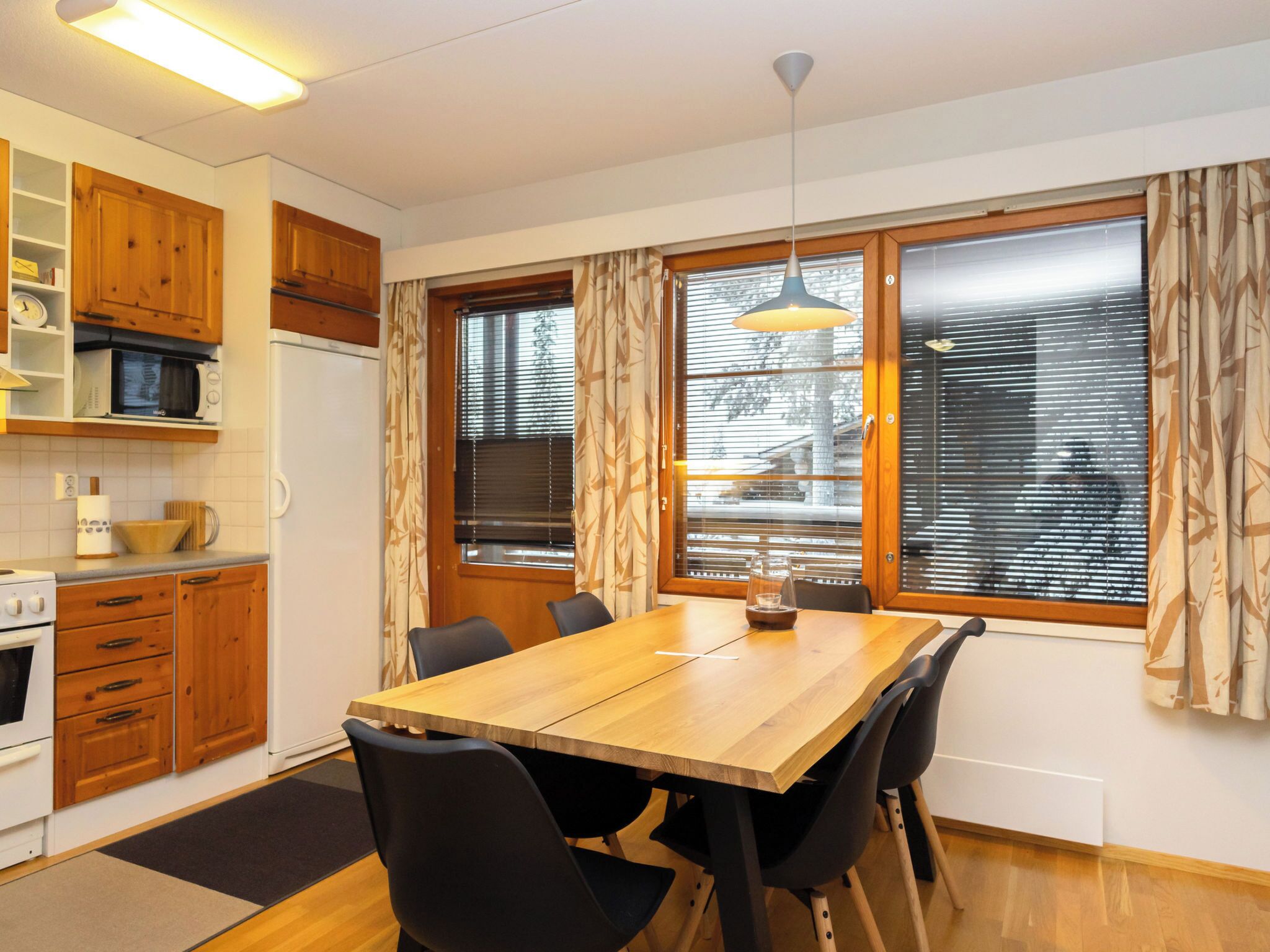Cocina compacta con armarios de madera, microondas y estufa. Hay una mesa de comedor con sillas y una lámpara colgante.