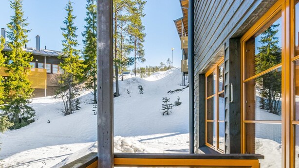 Una terraza de madera con un banco que da a un paisaje nevado con árboles y un edificio al fondo.