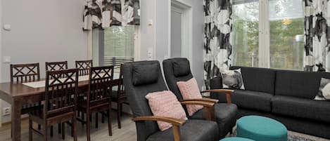 A modern living room with a dining area, grey sofas, wooden chairs, and a teal ottoman.