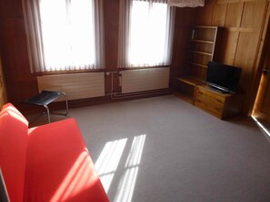 Ein Zimmer mit einem Bett, einem Fernseher auf einem Holzboden, einem Heizkörper und einem Fenster mit Vorhängen.