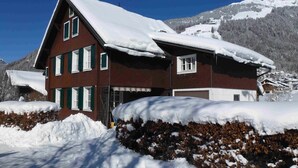 A multi-story building with a sloped roof, surrounded by snow-covered ground and trees, with a mountain in the background.