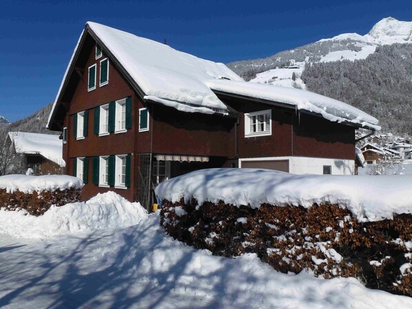 Um edifício de vários andares com telhado inclinado, rodeado por terreno e árvores cobertos de neve, com uma montanha ao fundo.