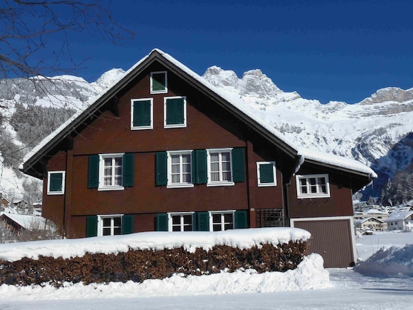 Un chalet de dos pisos con contraventanas verdes, techo cubierto de nieve y montañas al fondo.