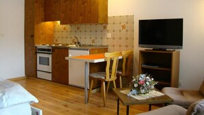 Ett kompakt kök med träskåp, en inbyggd ugn och en liten matplats med två stolar. En platt-TV är monterad på väggen ovanför en trähylla.