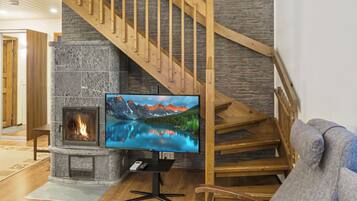 A living room with a stone fireplace, a television mounted on a stand, a wooden staircase, and a grey sofa.