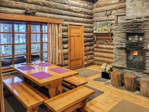 A log cabin interior with a wooden dining table, benches, a stone fireplace, and a large window overlooking a forest.
