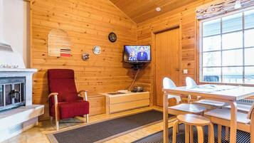 Tunnelmallinen mökin sisustus, jossa on puusisustus, seinälle asennettu televisio, punainen tuoli, ruokapöytä tuoleineen ja takka.