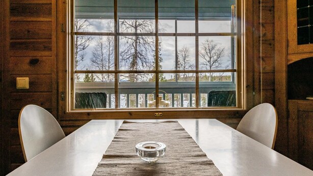 A long dining table with a candle centerpiece, wooden walls, and a view of trees through a large window.