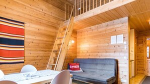 A cozy cabin interior with a wooden floor, a sofa, a dining table with chairs, and a ladder leading to a loft area.