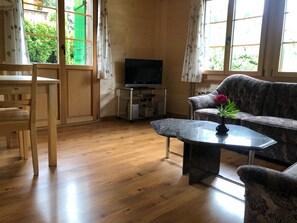 Una sala de estar con piso de madera, un sofá, una mesa de centro, un televisor y una ventana con cortinas.