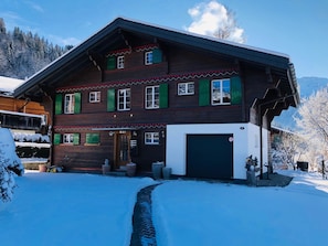 Una casa de madera de dos pisos con un garaje, contraventanas verdes y un camino de entrada cubierto de nieve.
