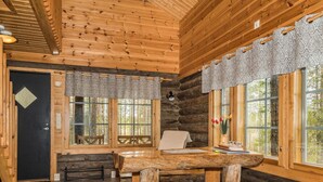 L’intérieur d’une cabane en rondins, avec une table à manger en bois, des bancs et de grandes fenêtres ornées de rideaux à motifs.
