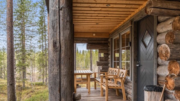 Une cabane en rondins avec une véranda en bois, une table et des chaises, et une porte ornée d’un motif en losanges.