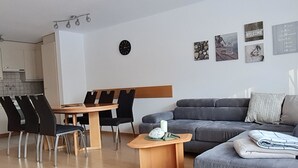 Uma sala de estar moderna com um sofá cinzento, uma mesa de centro de madeira e um espaço de jantar com mesa de madeira e cadeiras pretas.