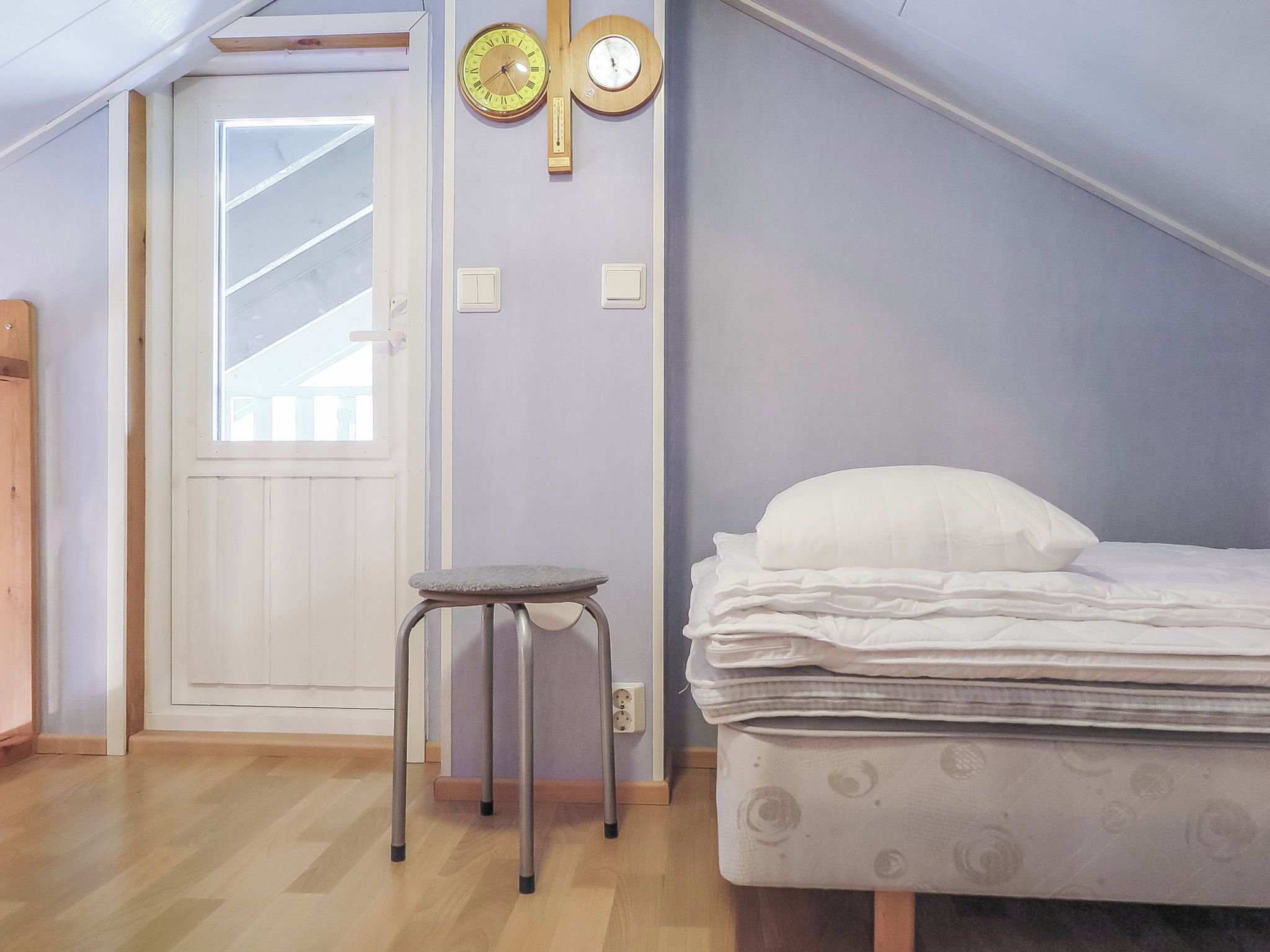 Quarto com uma cama, um banquinho pequeno, uma porta e dois relógios de parede.