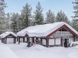 A wooden cabin with a snow-covered roof, surrounded by snow and trees.