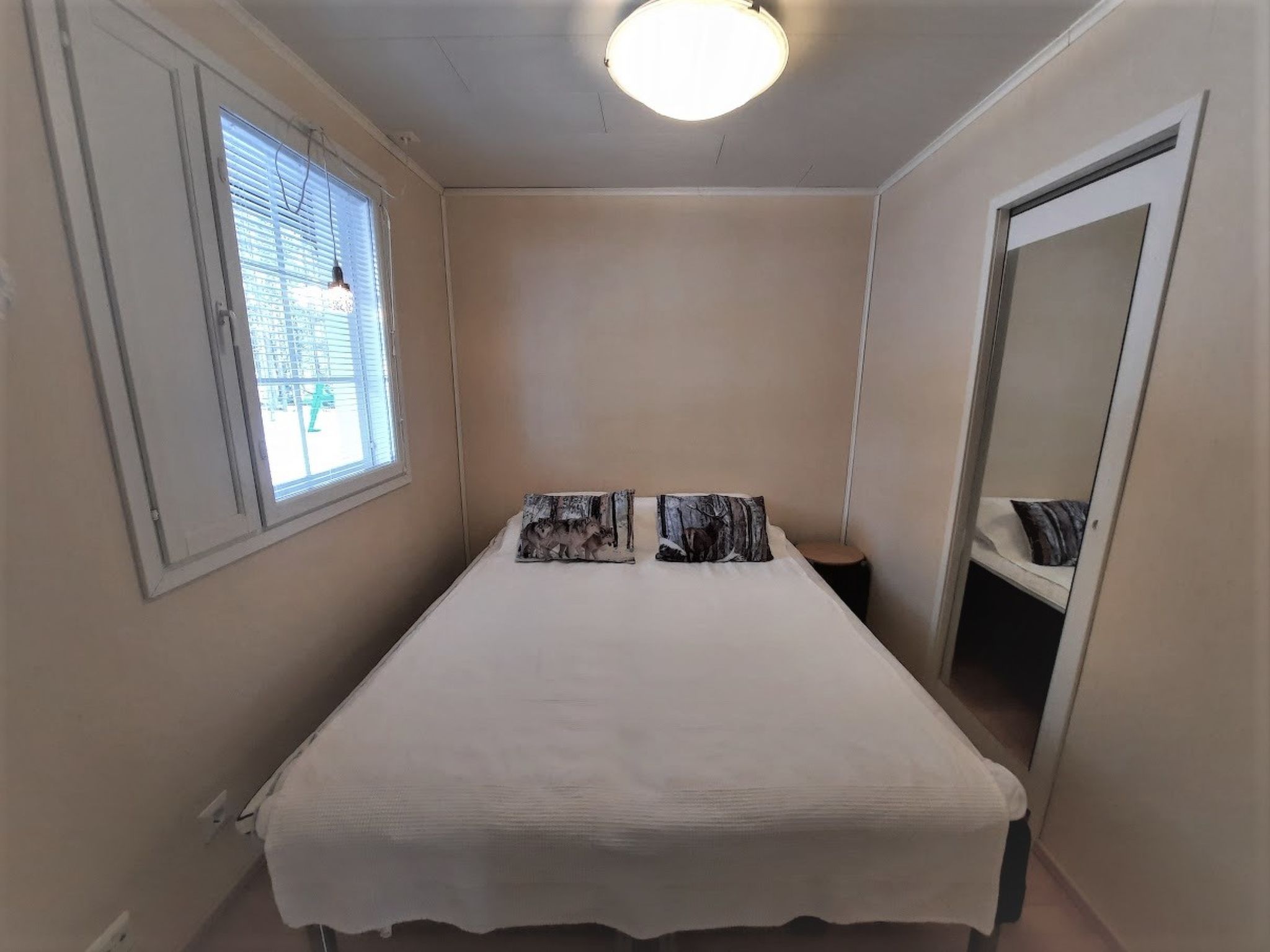 Quarto pequeno e estreito com uma cama de solteiro, janela com persianas, luminária no teto e espelho na parede.