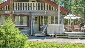 Un chalet rustique avec une façade en bois, un balcon doté de balustrades blanches et un petit espace porche aménagé avec des sièges d’extérieur.