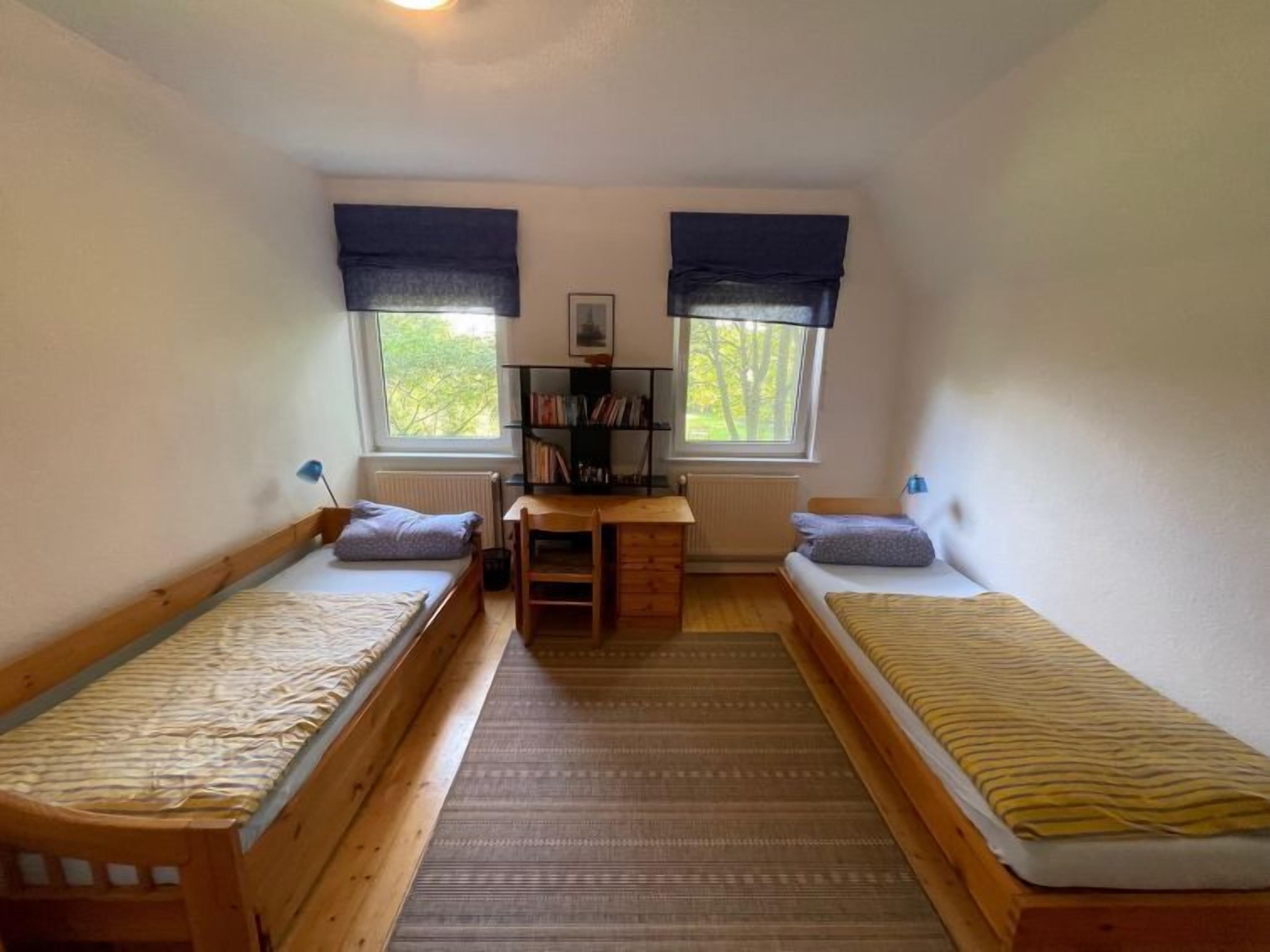 A small, well-lit room with two single beds, a desk with a chair, and a bookshelf.
