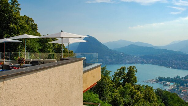 Een terras met buitenmeubels en parasols met uitzicht op een meer en bergen.