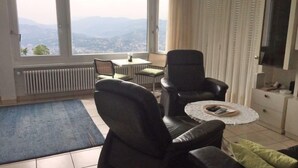 Een woonkamer met uitzicht op bergen, een zwarte leren bank, een klein rond tafeltje en een televisie aan de muur.