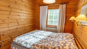 Ein Zimmer einer Holzhütte mit einem Bett, einem Fenster mit Vorhängen und einer Lampe.