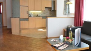 Een moderne keuken met houten kastjes en een ingebouwde oven. Er is een eethoek met een tafel en stoelen, en een woonkamer met een bank en salontafel.