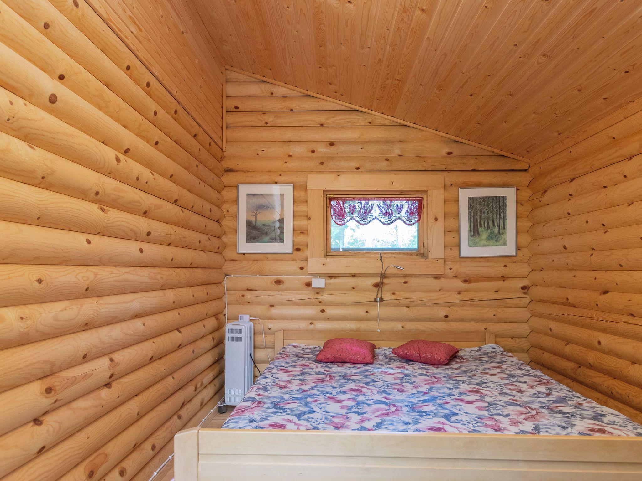 A small, cozy room with wooden walls and a sloped ceiling. There is a bed with a floral bedspread and two pillows, a window with a decorative curtain, and two framed pictures on the wall.