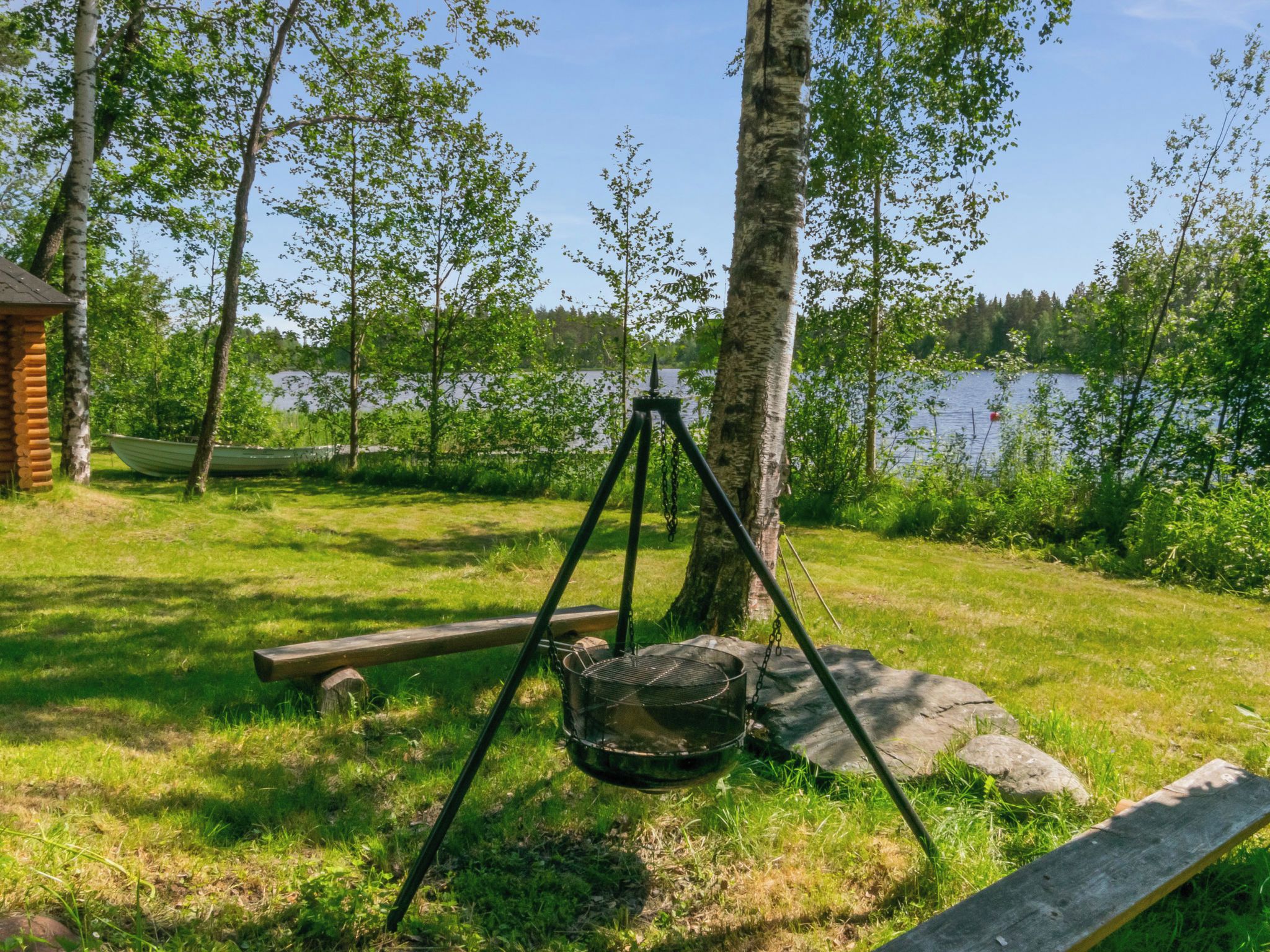 Une terrasse en bois avec un barbecue en métal, un banc en bois et une petite cabane située dans une zone boisée, avec un lac en arrière-plan.