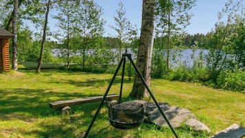 A wooden deck with a metal grill, a wooden bench, and a small cabin in a wooded area with a lake in the background.