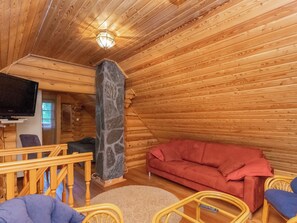 L’intérieur d’un chalet accueillant, avec une cheminée en pierre, un plafond en bois, un canapé rouge et un téléviseur posé sur un meuble en bois.