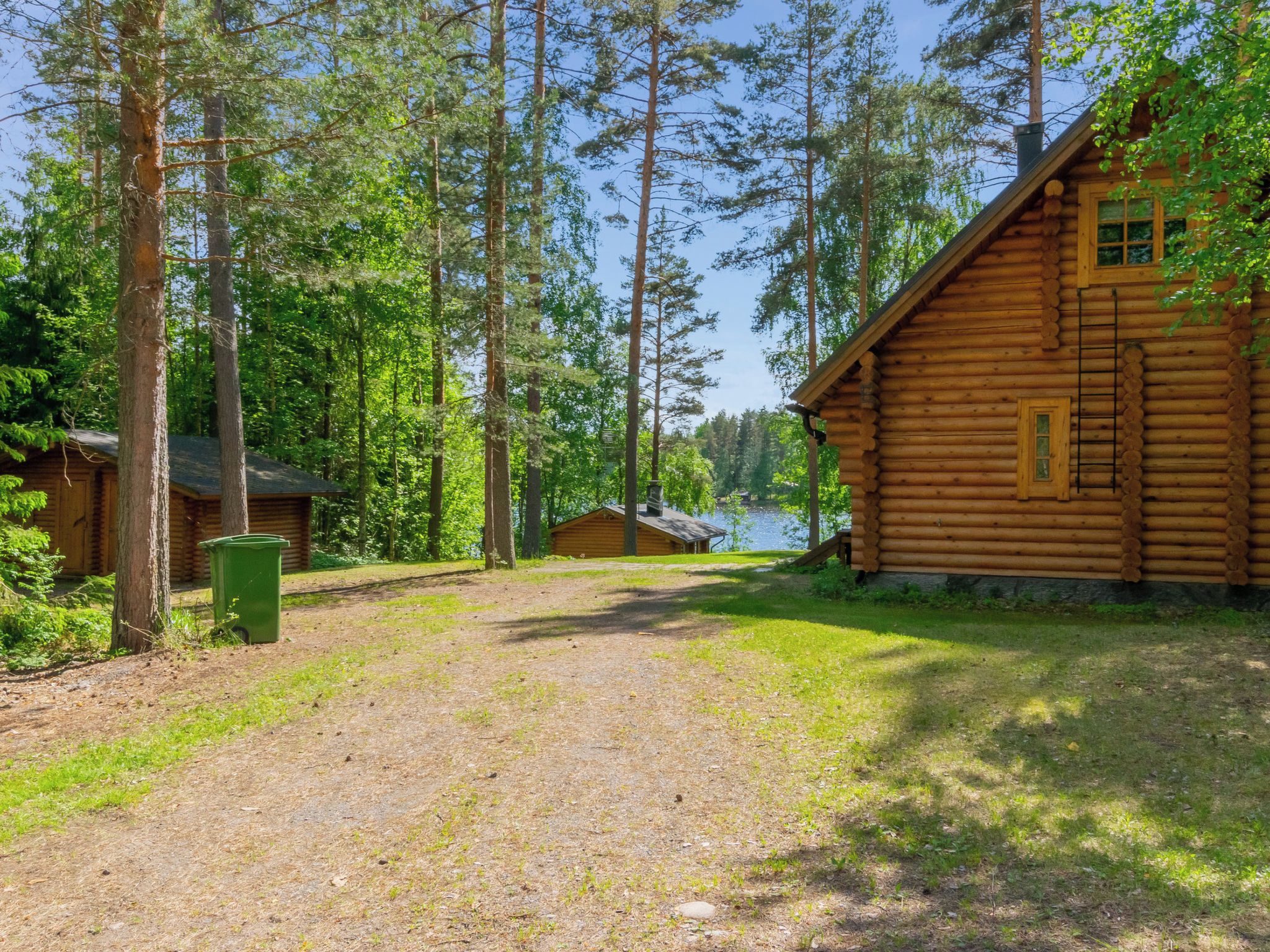 Une cabane en rondins, accessible par un chemin de gravier, entourée de grands arbres et avec une étendue d’eau en arrière-plan.