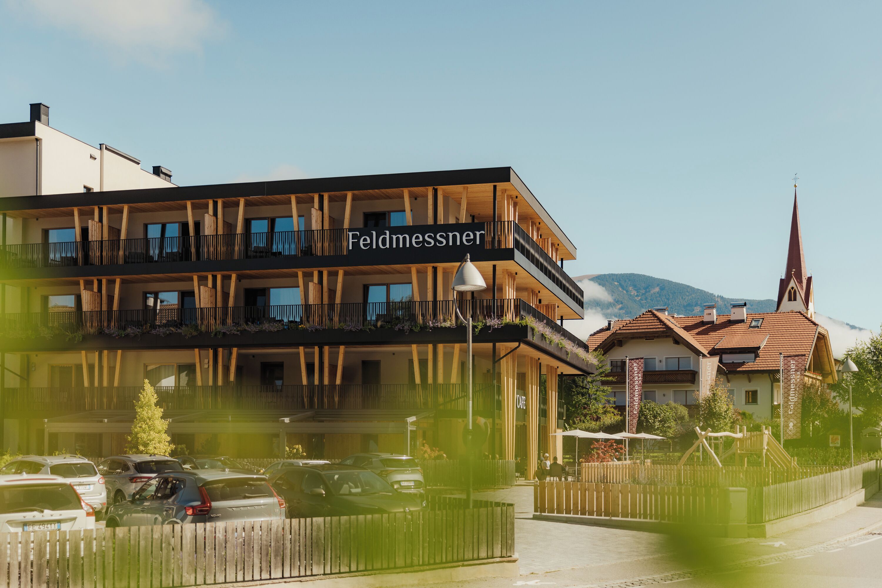 Ein Hotel mit Namenstafel, mehreren Balkonen und Blick auf eine Kirche und Berge.