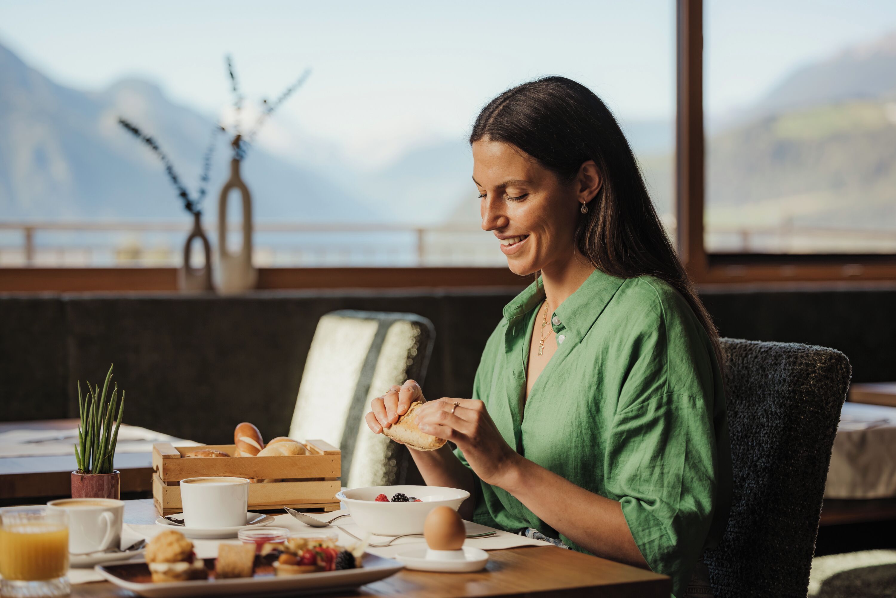 Une personne est assise à une table, devant un petit-déjeuner, avec une fenêtre donnant sur un paysage de montagnes et une plante en arrière-plan.