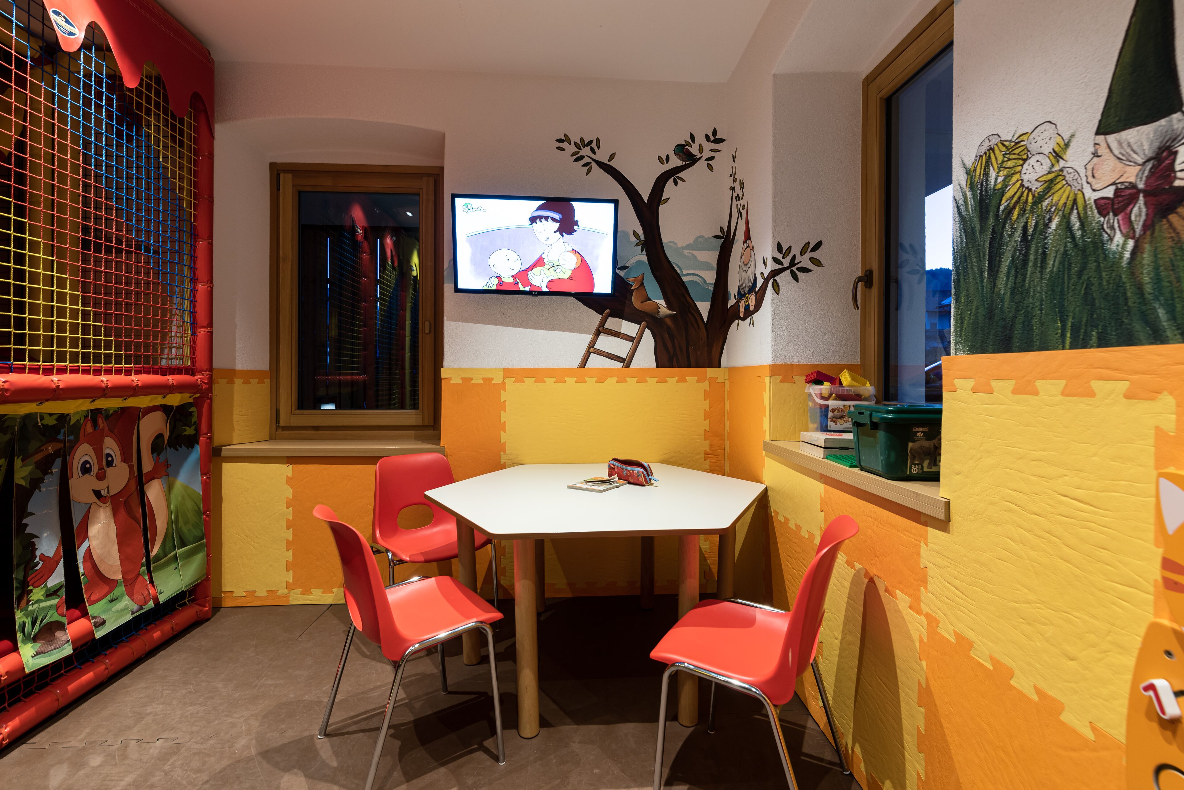Un área de juegos infantiles con televisor, sillas rojas y un mural con un árbol.