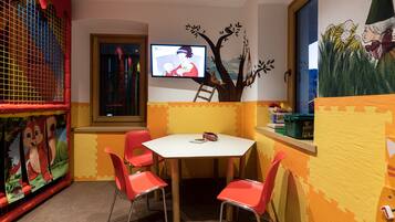 Un área de juegos infantiles con televisor, sillas rojas y un mural con un árbol.