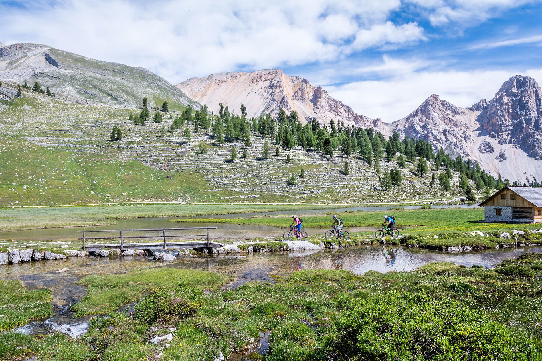 A mountain landscape with a small wooden cabin, a bridge over a stream, and cyclists on a trail.
